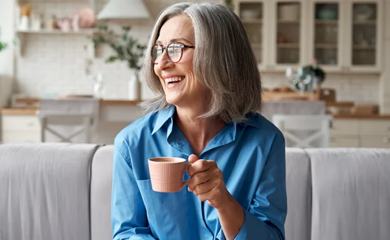 A happy and beautiful, relaxed, mature, grey-haired woman drinking coffee and relaxing on a sofa in a modern living room.